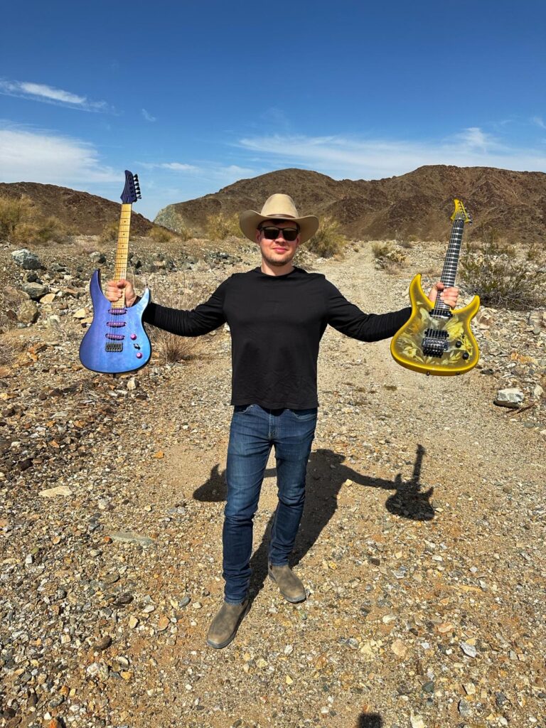 Rafal Perz Ruf Guitars Ceo holding two guitars on the desert. One blue, one gold.